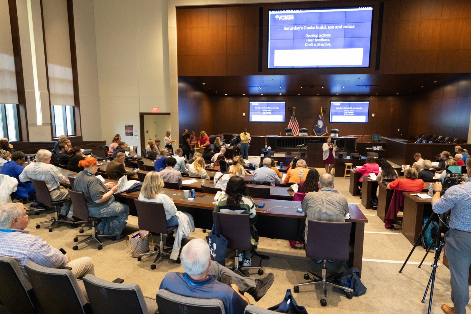Delegates seated in the assembly chamber during a plenary discussion, with "Saturday’s Goals: build, test and refine" on the screens